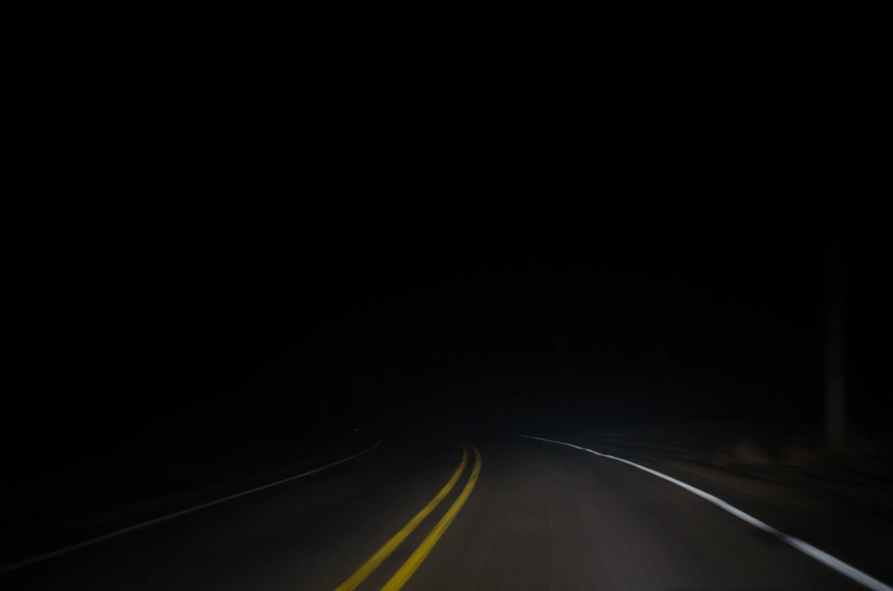 Dark road at night lit by headlights, yellow dividing lines stretching into the distance
